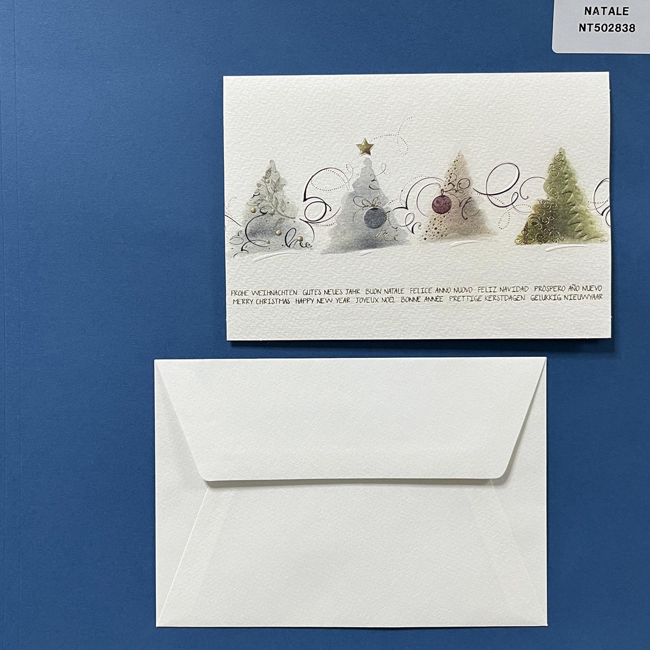 Elegante biglietto di Natale su carta martellata, con in copertina una simpatica illustrazione con piccoli alberi di Natale stilizzati e scritte augurali in più lingue, in oro a caldo.